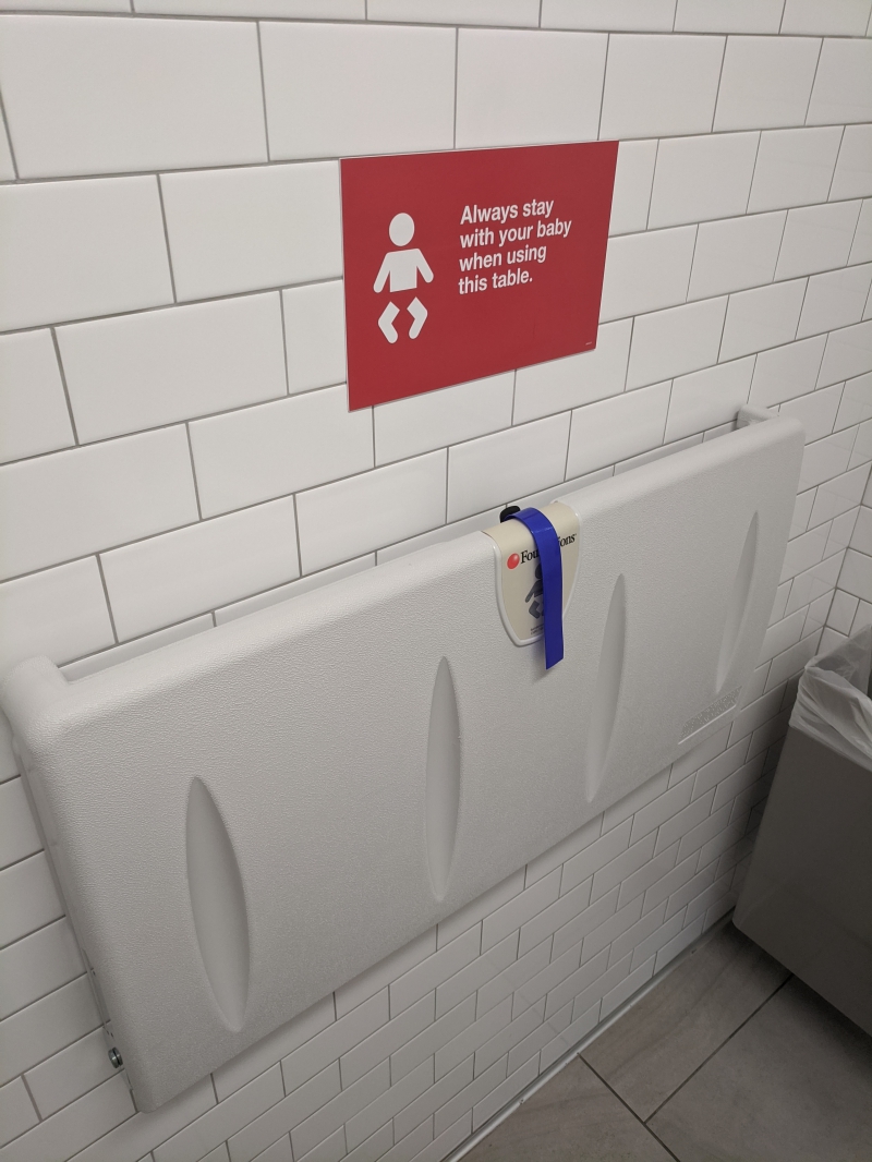 A white tiled wall with a red sign warning to stay with your baby. Below the sign is a white plastic changing table hooked to the wall. Photo by Tom Ackerman.