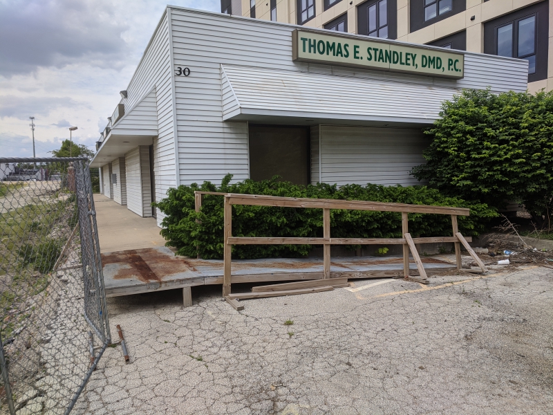 A wooden wheelchair ramp along the side of an office building with white siding. It leads to a sidewalk that wraps around the building. Photo by Tom Ackerman.