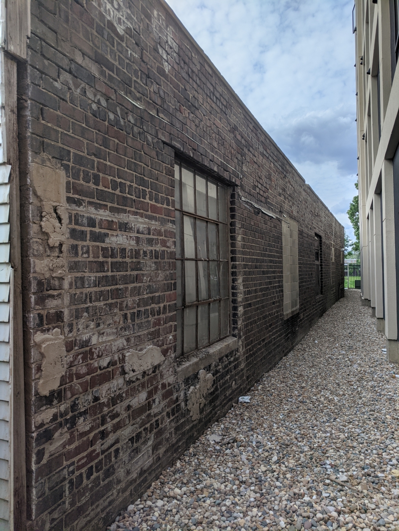 A narrow gravel alley in between a building with a brick wall and an apartment building. Photo by Tom Ackerman.