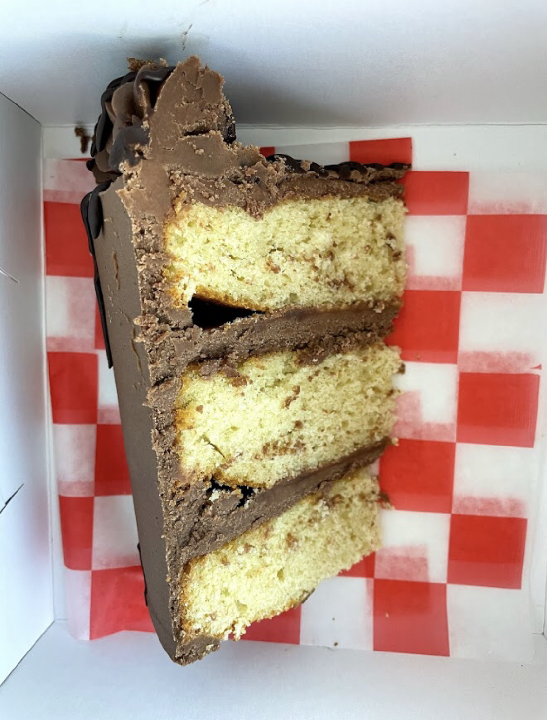 A three layer vanilla cake with light chocolate frosting sits in a white box on red and white checkered parchment paper. Photo by Remington Rock.