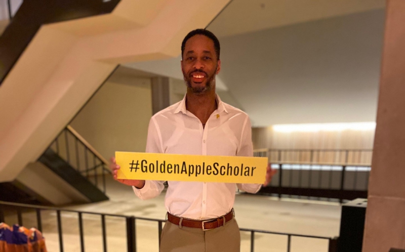 A Black man with short hair and a beard is holding a yellow sign that says #GoldenAppleScholar in black letters. He is wearing a white collared shirt. Photo provided by Mark Butler.