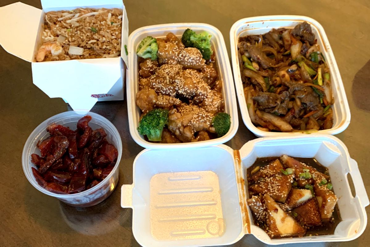 A photo looking down on a spread of (clockwise) to-go containers filled with sesame chicken covered in sesame seeds, Mongolian beef with mixed vegetables in a thin sauce, crispy tofu in a dark, sticky sauce and covered in sesame seeds, thinly sliced beef rib pieces covered in a dark red honey sauce and a carton filled to the top of shrimp fried rice with pieces of onion, been sprout and shrimp visible at the top. Photo by Megan Friend.