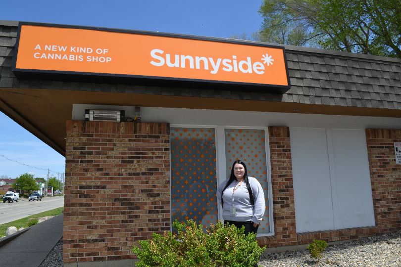 Nikki Haese, dispensary owner, stands in front of the exterior of Sunnyside Champaign is a small brick building with a bright orange sign reading Sunnyside. Photo by Alyssa Buckley.