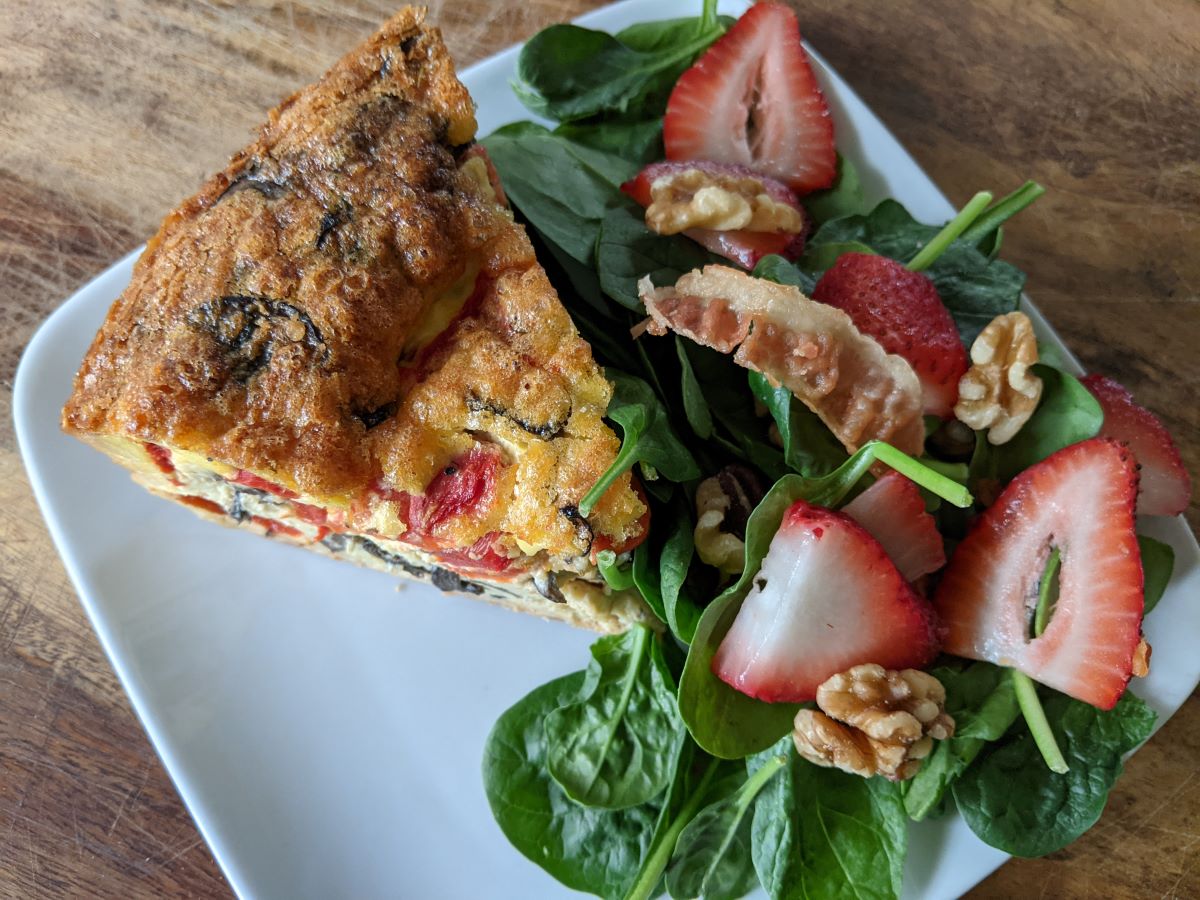 A slice of golden quiche with a green spinach salad topped with strawberries on the side.  The food is set on a square, white plate that sits on a block of wood. Photo by Tias Paul.
