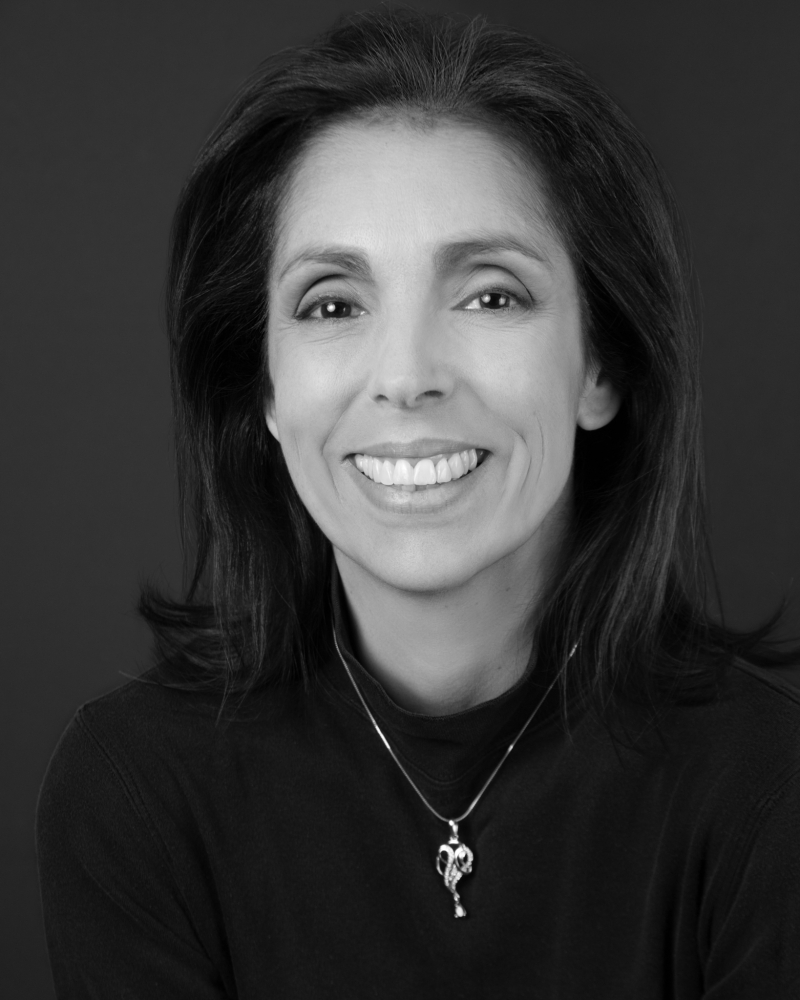 Black and white headshot of smiling woman with dark eyes, shoulder length dark hair, black shirt, and silver necklace. Photo from Luciana Rezende-Bagby.