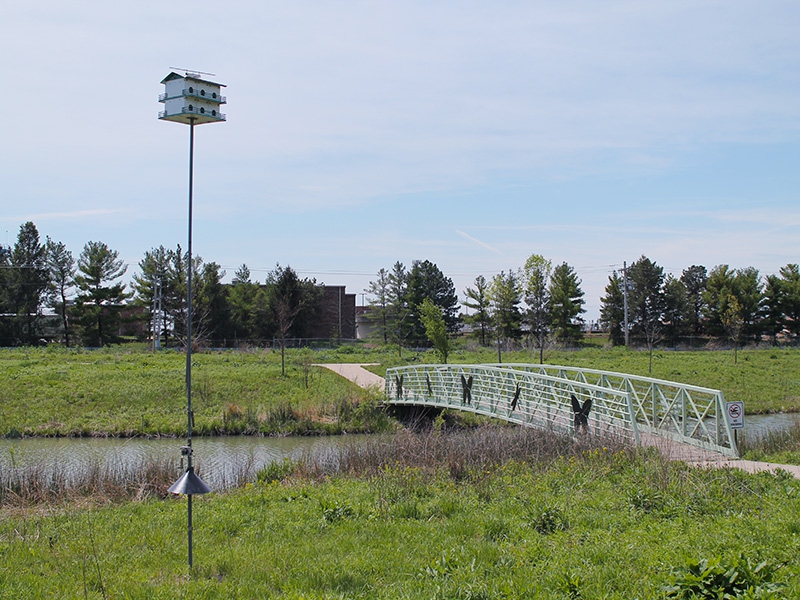 a small pedestrian bridge over a pond with a tall birdhouse in the foreground