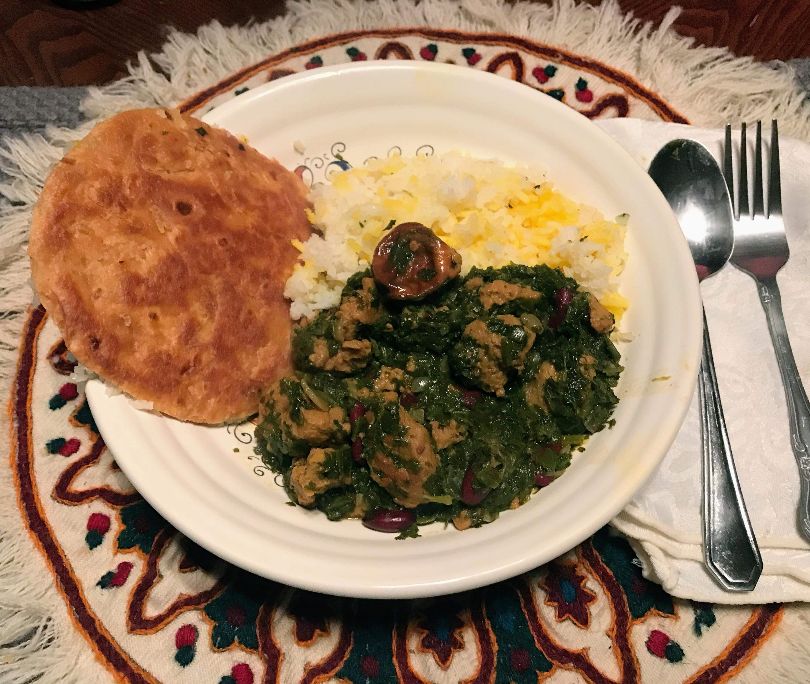 On a table with a circular, fringed placemat, there is a white plate with an Iranian dish called Ghorem Sabzi with Persian rice. Photo by Darya Shahgheibi.