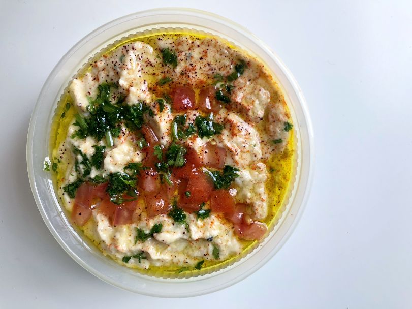 An overhead photo of baba ghanoush shows a plastic takeout bowl filled with the eggplant dip topped with seasoning, herbs, and diced tomato. Photo by Alyssa Buckley.