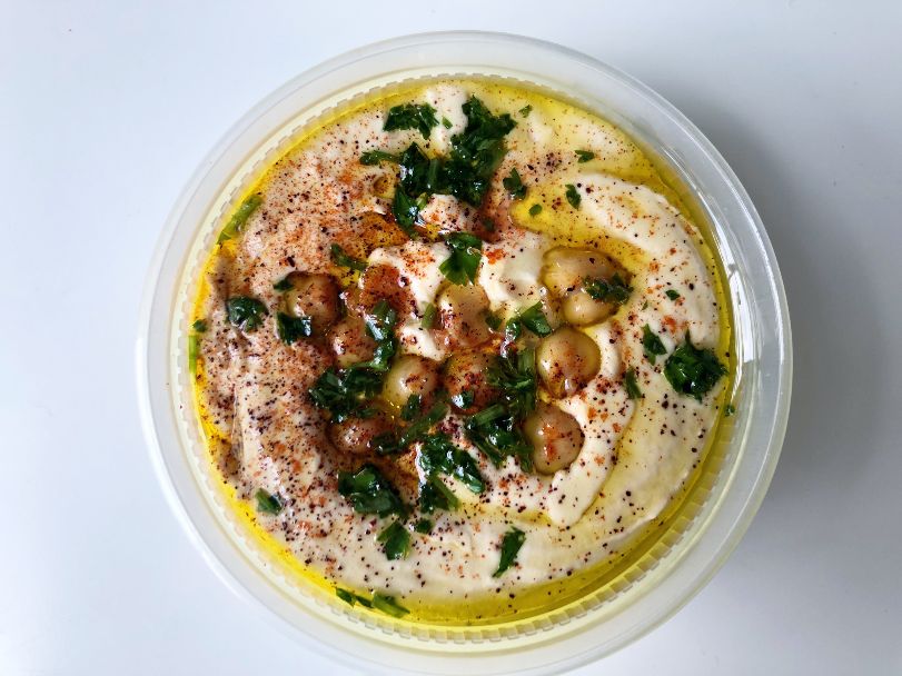 An overhead photo of hummus shows a circle takeout container with hummus topped with herbs, seasoning, and chickpeas. Photo by Alyssa Buckley.