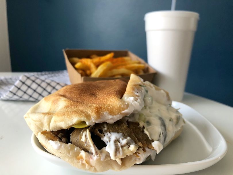 A pita from Shwarma Joint its on a white plate on a white table. The pita is slightly open and white sauce is covering the top layer and spilling out. The gyro meat is shaved thin and stick out a little on the left. In the background, there is a side of fries and soda. Photo by Alyssa Buckley.