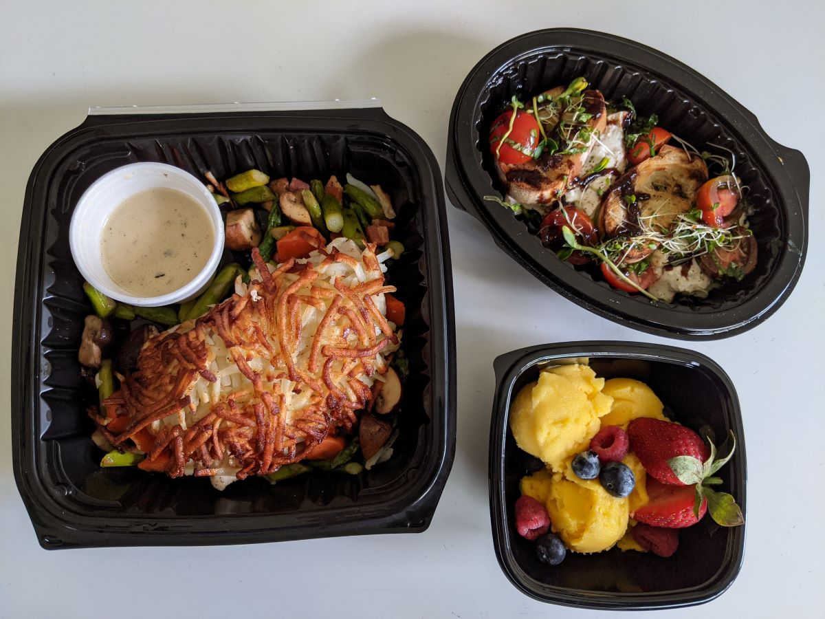 In the picture, there are three black, plastic containers of food sitting on a white tabletop.  On the left is a square container with potato-crusted fish, a round container of cream sauce, and colorful roasted vegetables. On the right, there is an oval container on the top of the picture contained the crostini, white cheese, red tomatoes, and green microgreens. Below the oval container is a small square container with orange mango sorbet, red strawberries, and blueberries. Photo by Tias Paul.
