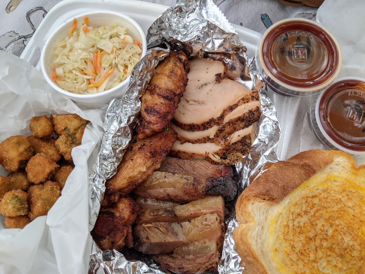 In the center of the picture from top to bottom are the sliced meats and wings. A container of slaw is at the top left corner of the picture and breaded fried okra is below it.  On the right side of the picture, two containers of BBQ sauce are at the top corner and a slice of golden toast is below it. Photo by Tias Paul.