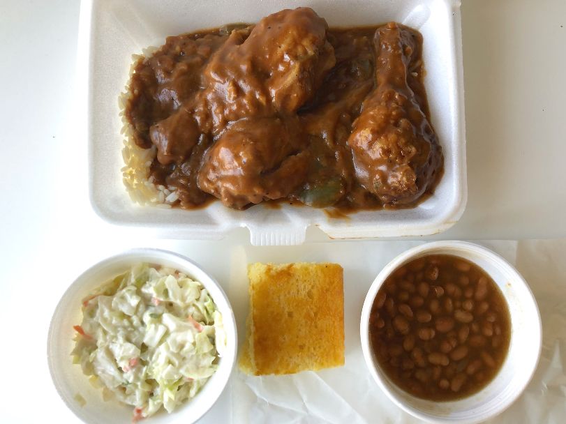 An overhead photo shows three chicken legs covered in gravy over rice in a styrofoam takeout container. There are three sides below: a white bowl of creamy coleslaw, a square of cornbread on white parchment paper, and a bowl of beans. Photo by Alyssa Buckley.