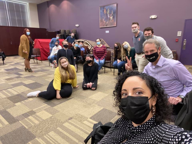 A group of people are gathered in a room, some standing some sitting in chairs, some on the floor. They are all wearing masks. Photo from Organic Oneness UIUC Facebook page.