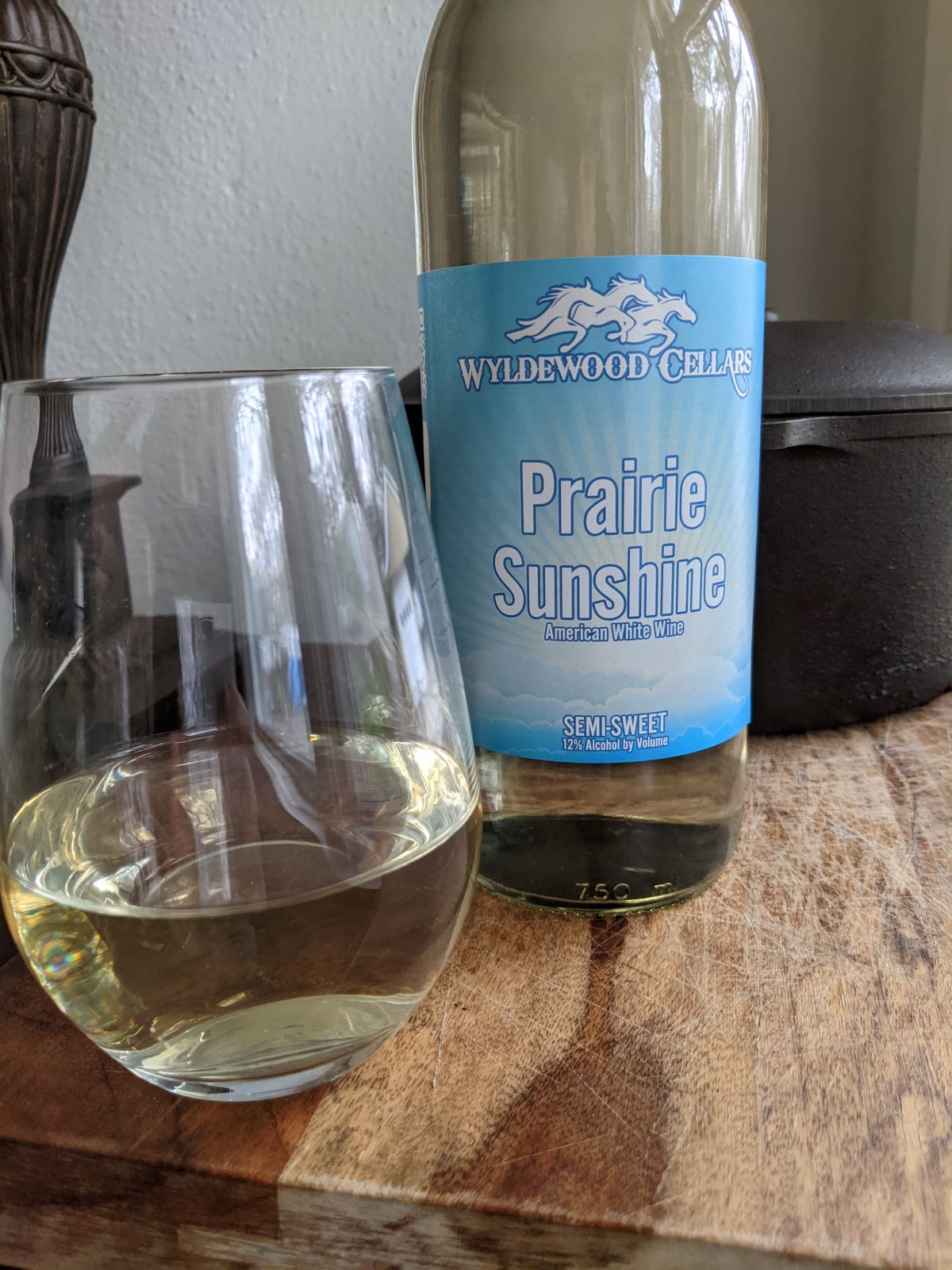 In the forefront of the picture is a bottle of white wine with a blue â€œPrairie Sunshineâ€ label and a glass of wine.  The glass and bottle sit atop a wooden block.  Behind these, we can see a black cast iron dutch oven and a bronze lamp. Photo by Tias Paul.