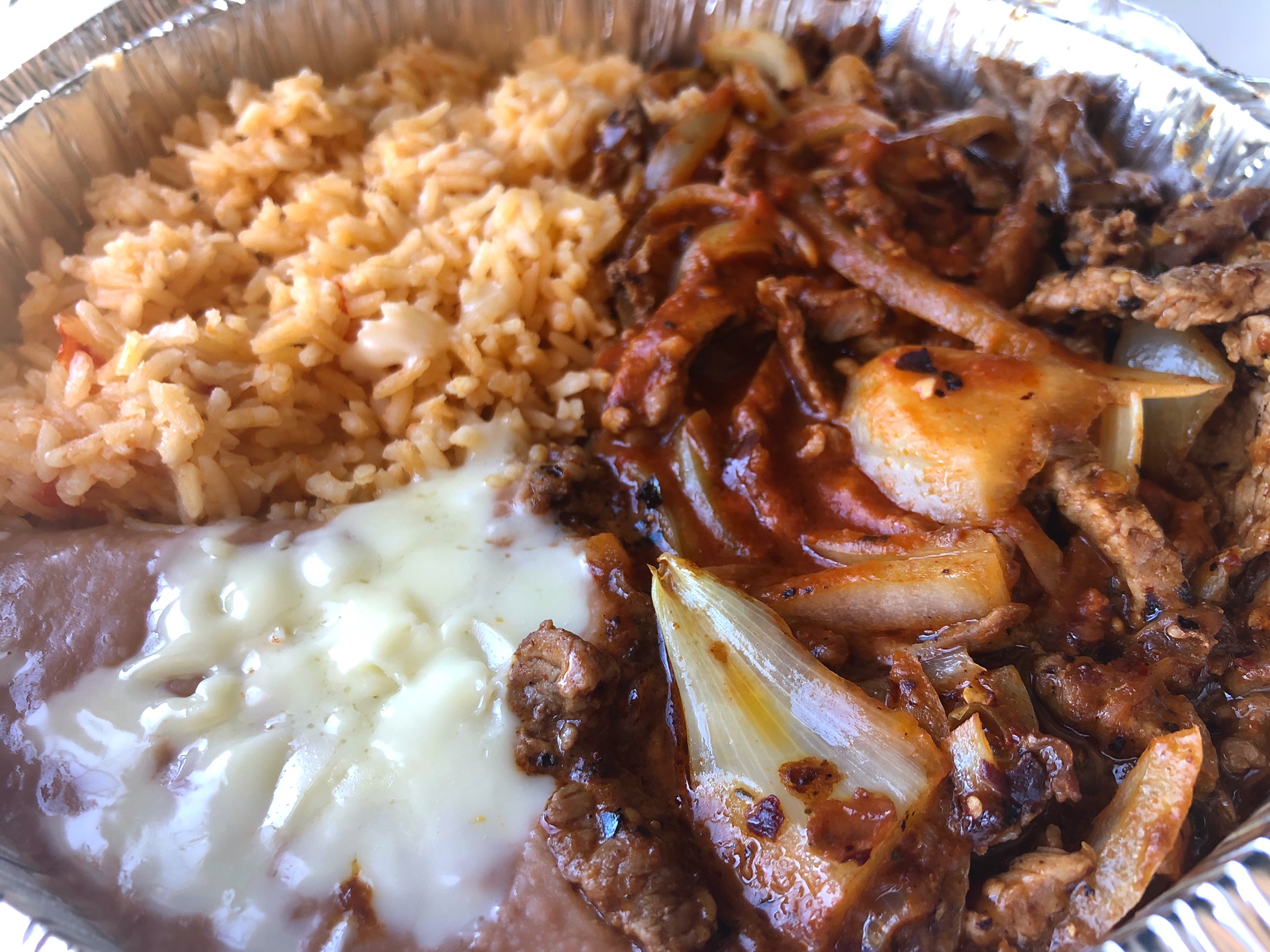In a tin circular takeout container, there is an ample portion of orange Mexican rice, then a dark red sauce covered beef Chile Colorado, and thin gray pinto beans with white melted cheese on top. Photo by Alyssa Buckley.