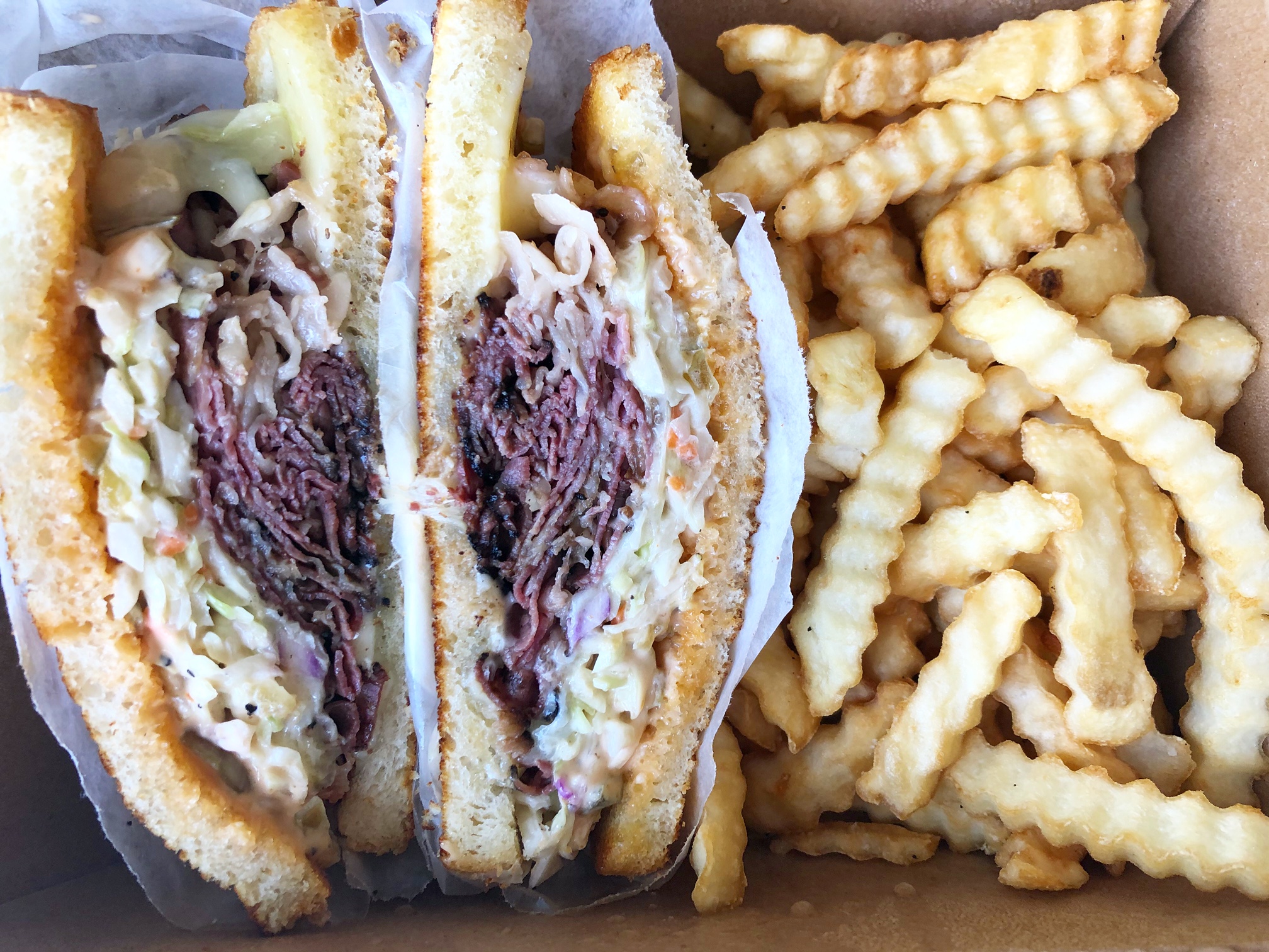 A pastrami sandwich is sliced in half and wrapped in parchment paper beside crinkle cut fries in a brown cardboard takeout box. Photo by Alyssa Buckley.