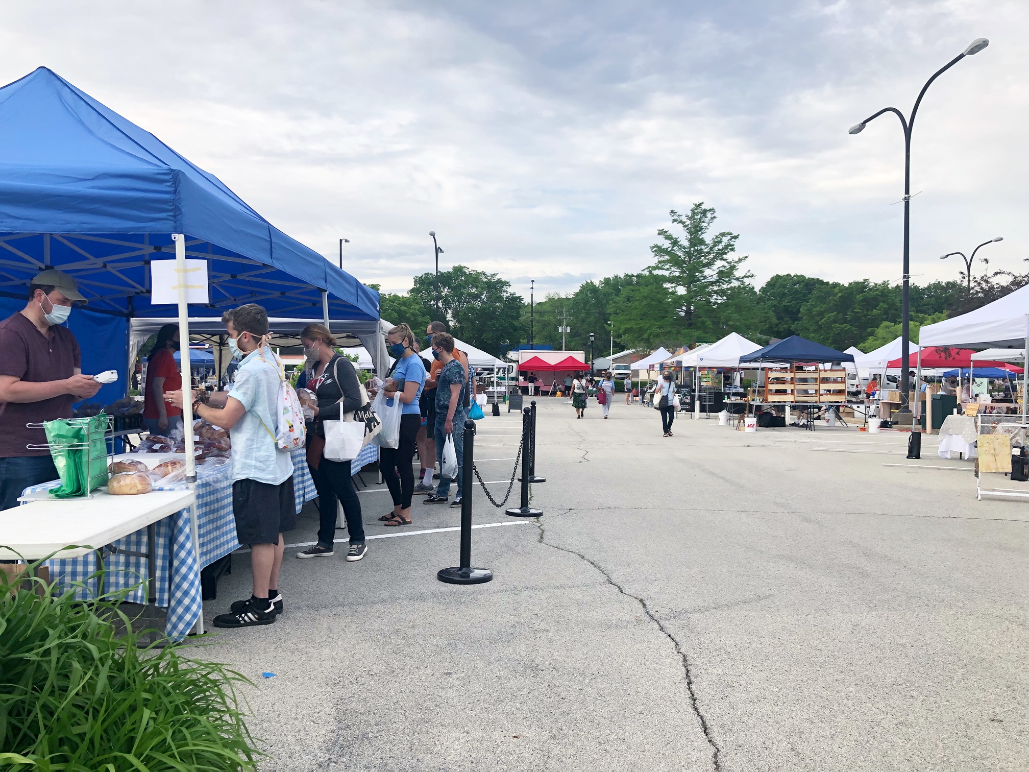 The Urbana market has shoppers outdoors in the Lincoln Square Mall parking lot. Vendors have blue or white tents with tables of goods for sale. Photo by Alyssa Buckley.