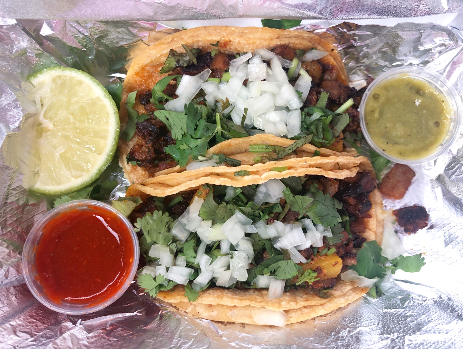 In a tin foil lined takeout container, there are two al pastor tacos, one lime wedge, a small cup of red salsa, and a small cup of green salsa. Photo by Alyssa Buckley.