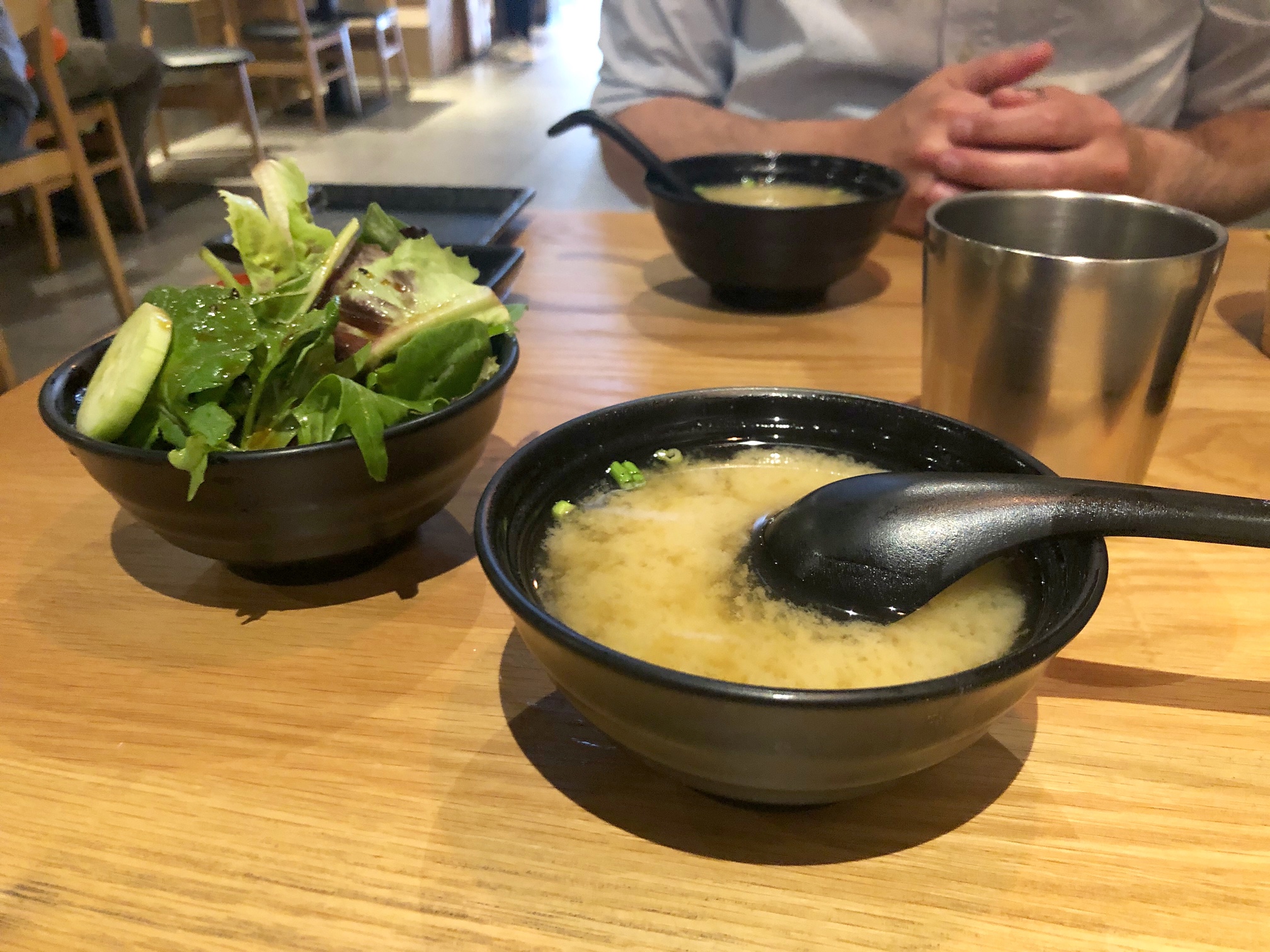 On a wooden table at Sakanaya, there is a black bowl filled with miso soup and a black spoon in front of a black bowl overflowing with salad greens. Photo by Alyssa Buckley.