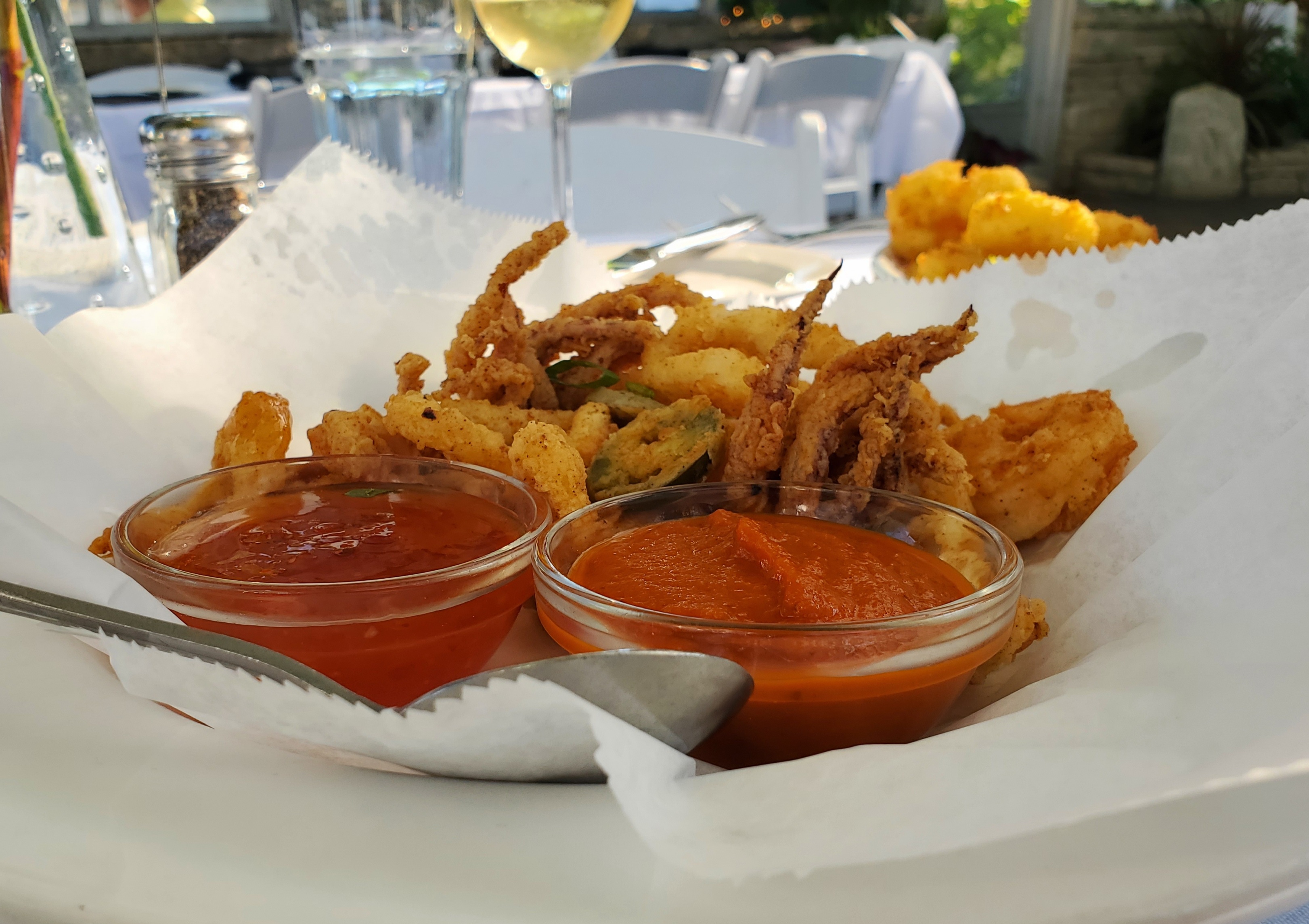 Fried shrimp and calamari are in a basket lined with parchment paper on a white table. In the basket, there are two small metal cups of dipping sauces. Photo by Carl Busch.