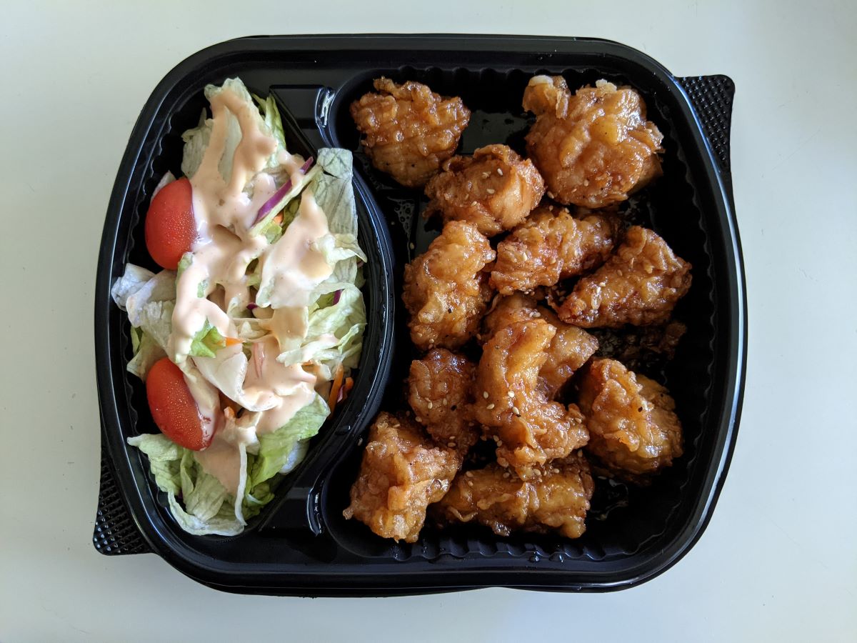 Fried chicken pieces coated in a golden crust with a shiny sauce in the large compartment of a two-compartment tray. The second (smaller) compartment features a tomato and lettuce salad with a pink dressing. Photo by Tias Paul.