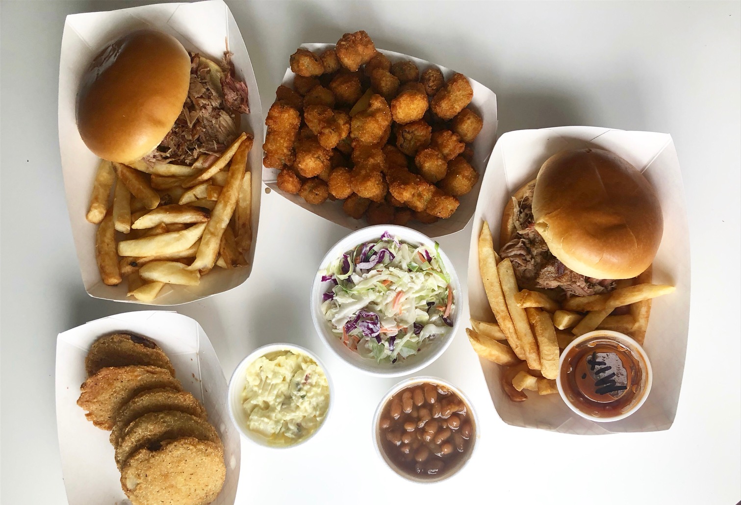 An overhead photo of the author's dinner with two pulled pork sandwiches, a basket of fried okra balls, a big cup of coleslaw, a small cup of potato salad, a small cup of baked beans, and a basket of fried green tomatoes. Photo by Alyssa Buckley.