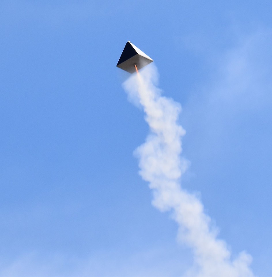 A pyramid shaped model rocket soars across a blue sky with a gout of orange engine fire leaving a trail of smoke in its wake. Photo by Christopher Brian Deem