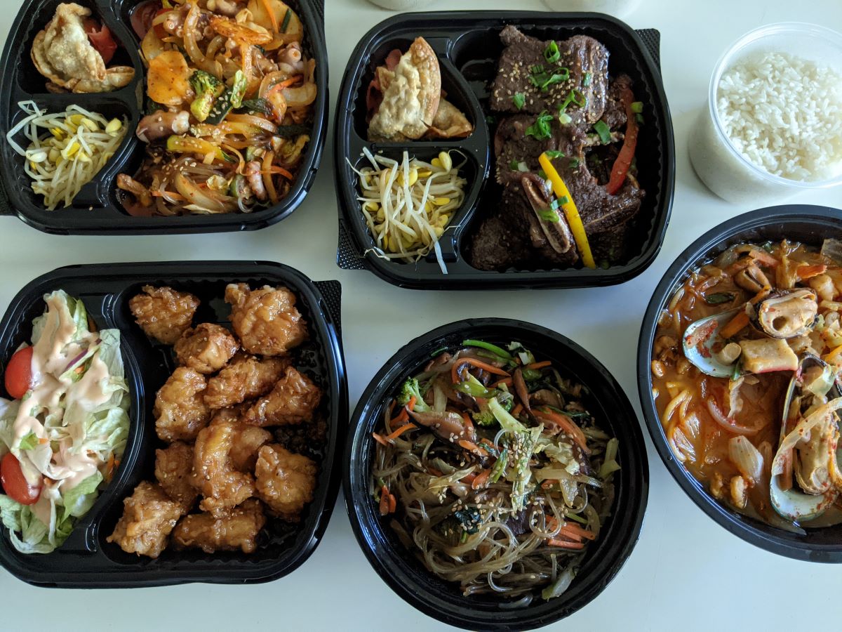 A top-down view of five entrees in black plastic trays plus rice in a clear plastic container placed on a white table. Photo by Tias Paul.