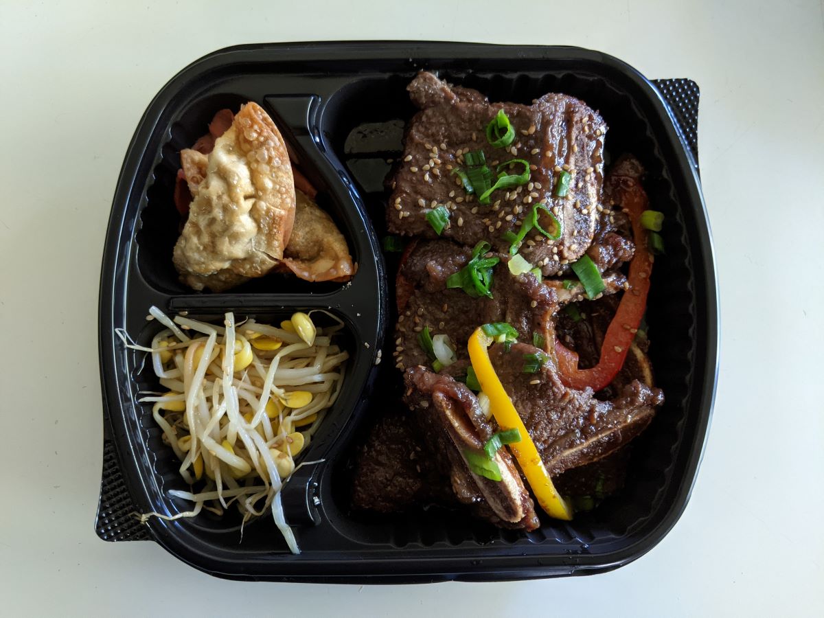 Brown beef short ribs with green onions and white sesame seeds on top in a black, three-compartment tray. Other compartments feature dumplings and sprouts. The tray is on a white table. Photo by Tias Paul.