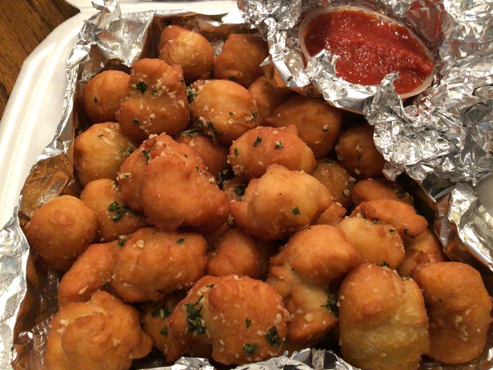 A white styrofoam takeout container lined with foil holds golden brown fried garlic butter doughnuts, which resemble small fritters. They are topped with a sprinkle of parmesan cheese and chopped fresh parsley. A plastic ramekin holding bright red pizza sauce for dipping sits in the corner of the dish. Photo by Rachael McMillan.