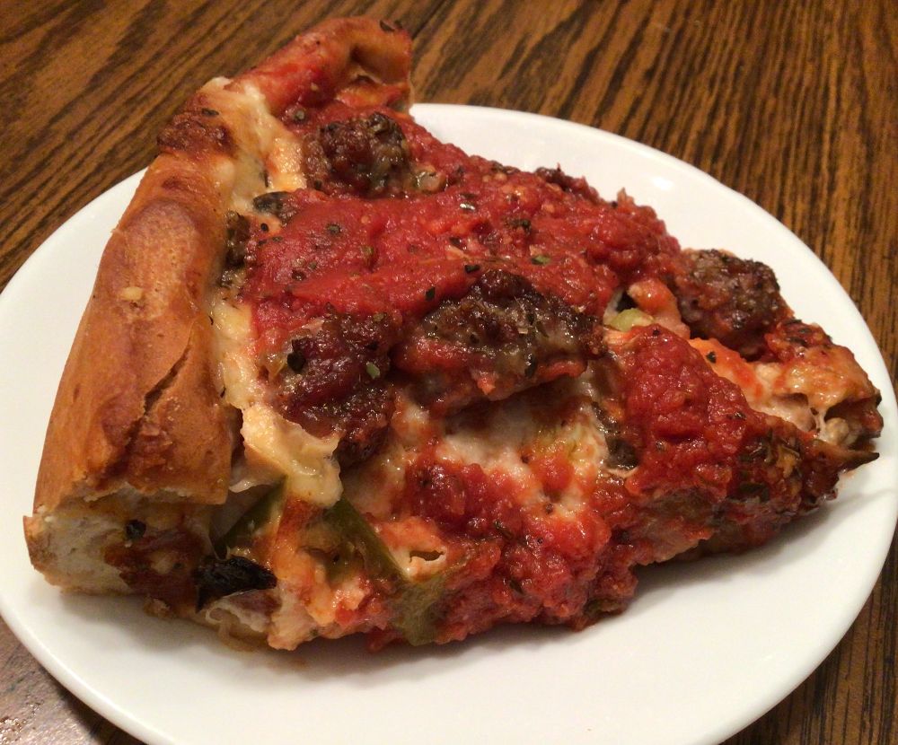 A small white plate holds a slice of deep dish pizza, shown slightly from the side. The toppings--sausage, mushroom, and green pepper, are all visible. The pie had been topped with a hearty red sauce before being sliced. Photo by Rachael McMillan.