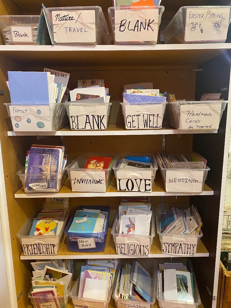 A set of shelves with plastic bins lining each shelf. The bins are filled with greeting cards, and have white signs in front labeling the style of card. Photo by Julie McClure.