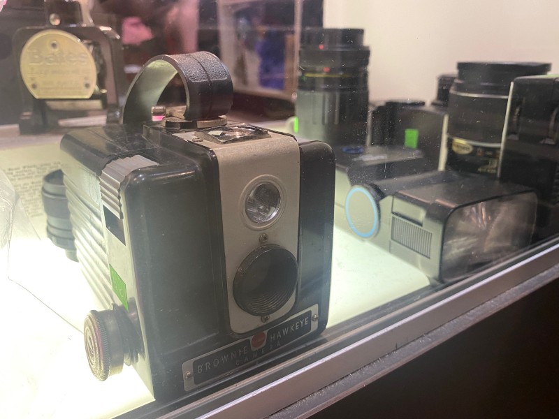 A cube shaped Brownie camera sits in a glass case. It is surrounded by other photography equipment. Photo by Julie McClure.