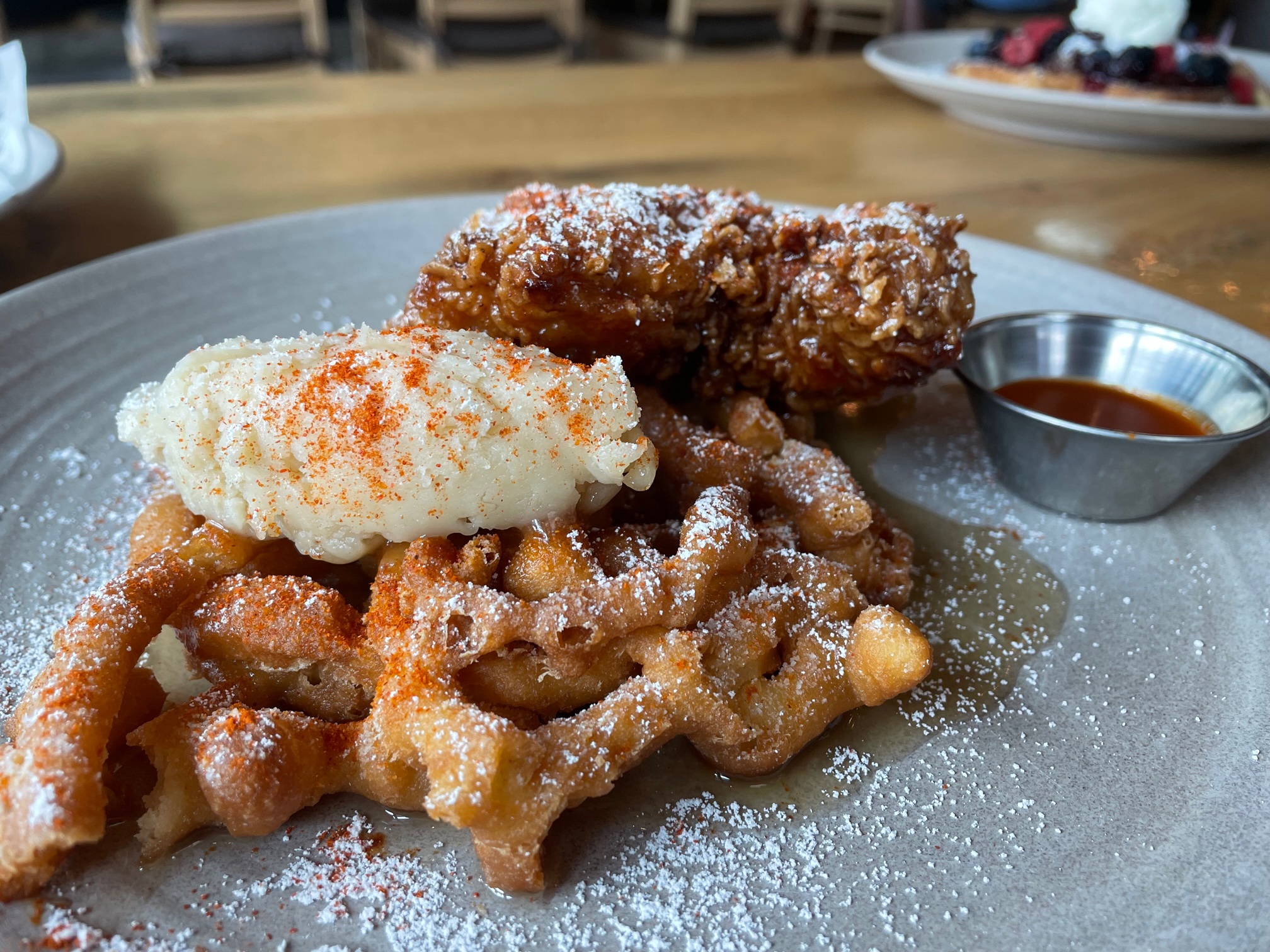 On a gray plate, there is a funnel cake with a large dollop of butter dusted with cayenne pepper beside Korean fried chicken dusted with powdered sugar. Photo by Alyssa Buckley.
