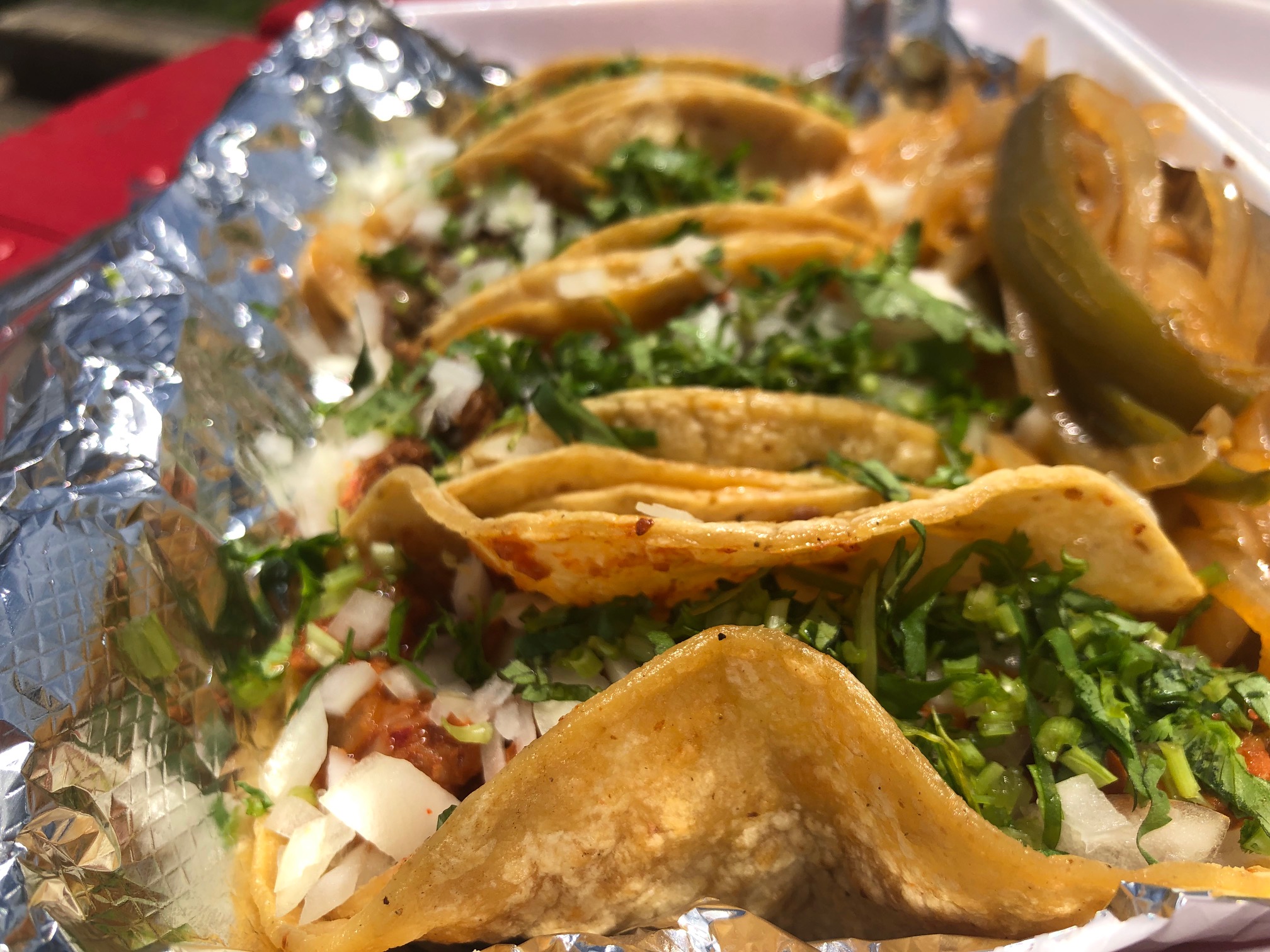 Four tacos on corn tortillas loaded with raw diced white onion and cilantro sit in a tin foiled takeout container. Photo by Alyssa Buckley.