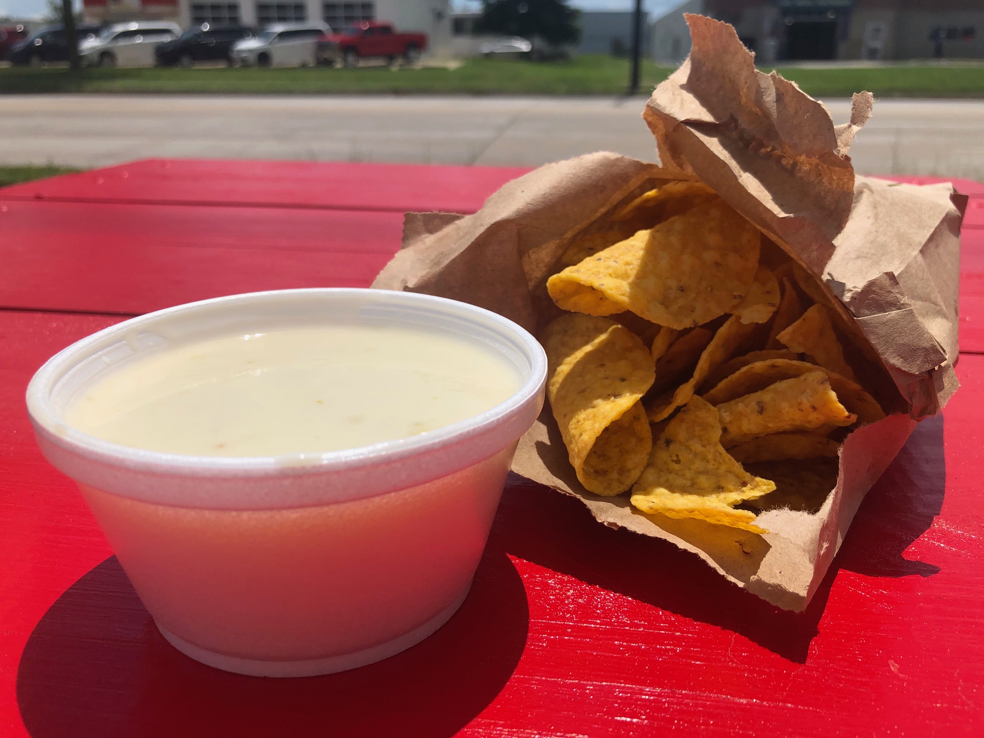 In a styrofoam cup, there is a white cheese dip, and beside that, there is a brown paper bag of orange chips on a red picnic table outside SeÃ±ores Taco in Champaign. Photo by Alyssa Buckley.