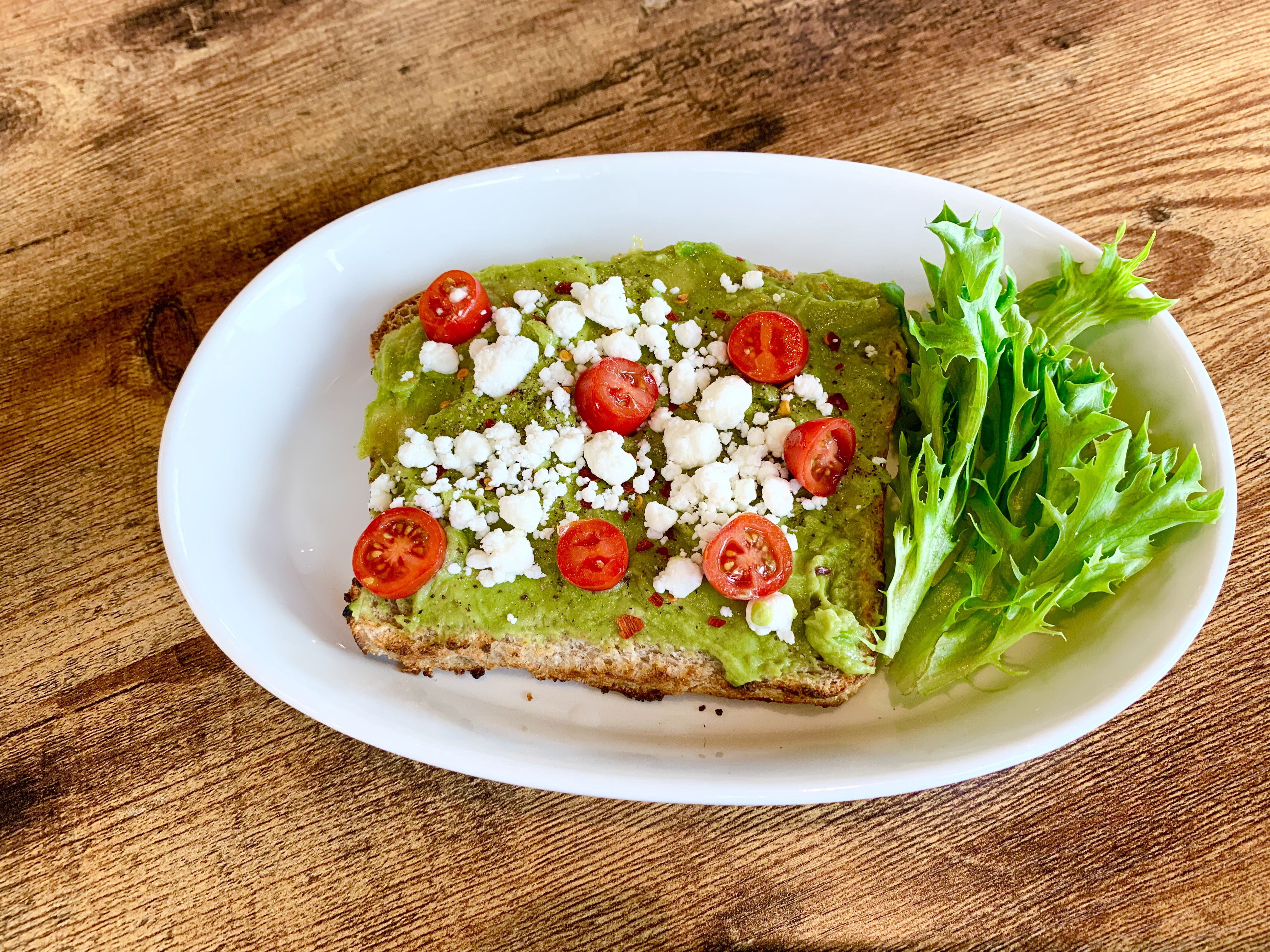 On a white plate, there is avocado toast topped with crumbled feta and tomatoes. Photo by Stephanie Wheatley.