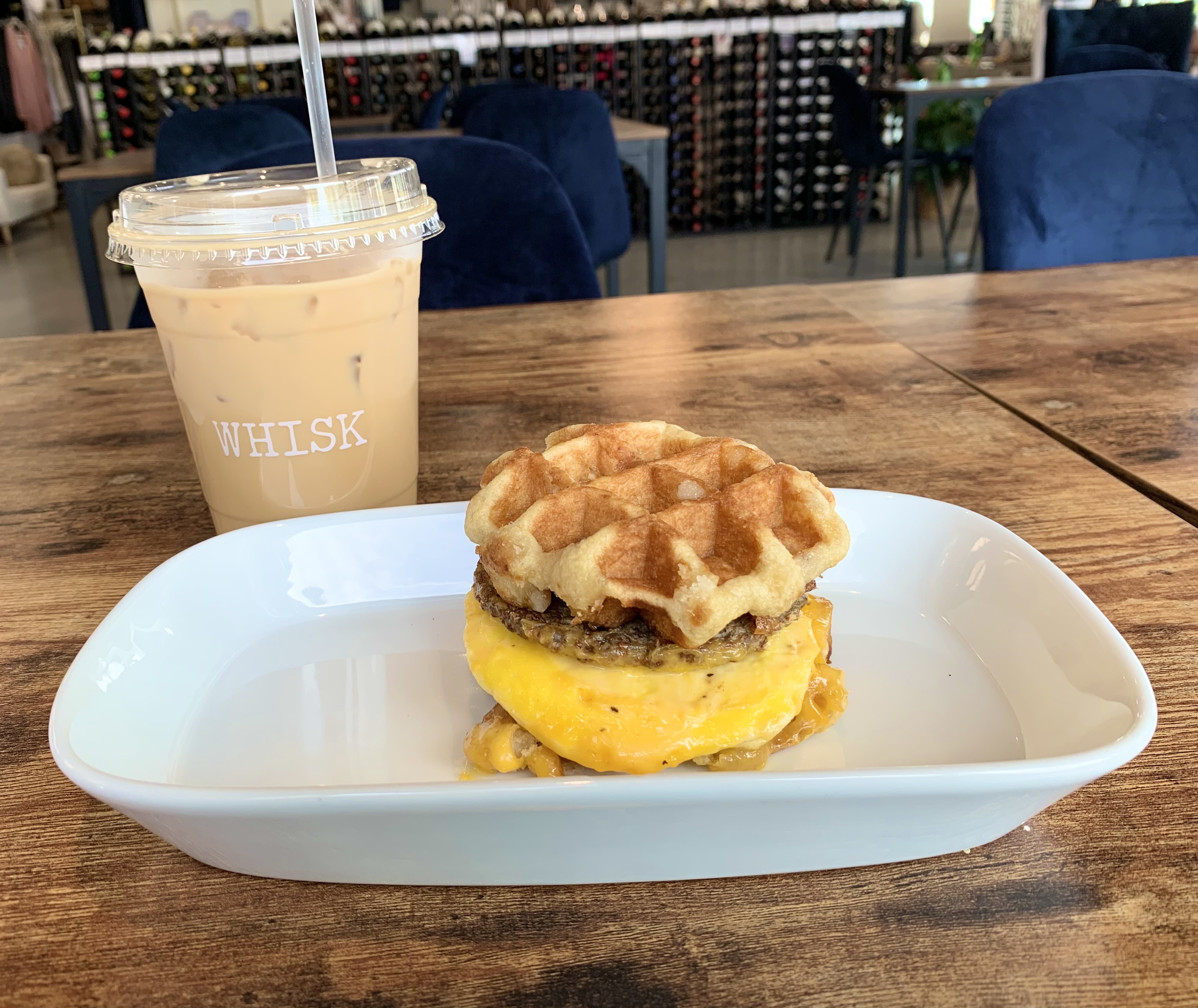 On a whiny white plate, there is a waffle breakfast sandwich. In the background, there is a brown coffee drink in a plastic cup. Photo by Stephanie Wheatley.