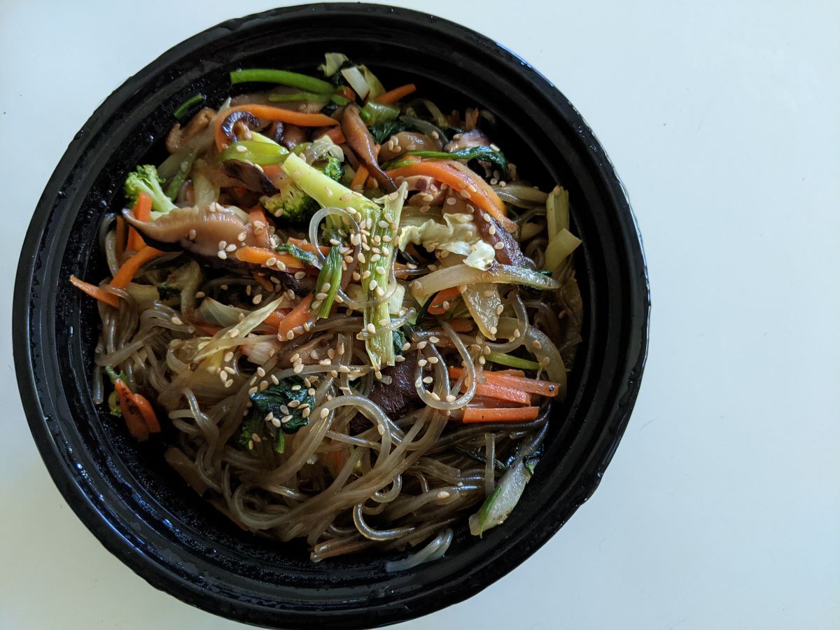 Light brown clear noodles with orange matchstick carrots, green onions, and white sesame seeds. The noodles are in a round, black container sitting atop a white table. Photo by Tias Paul.