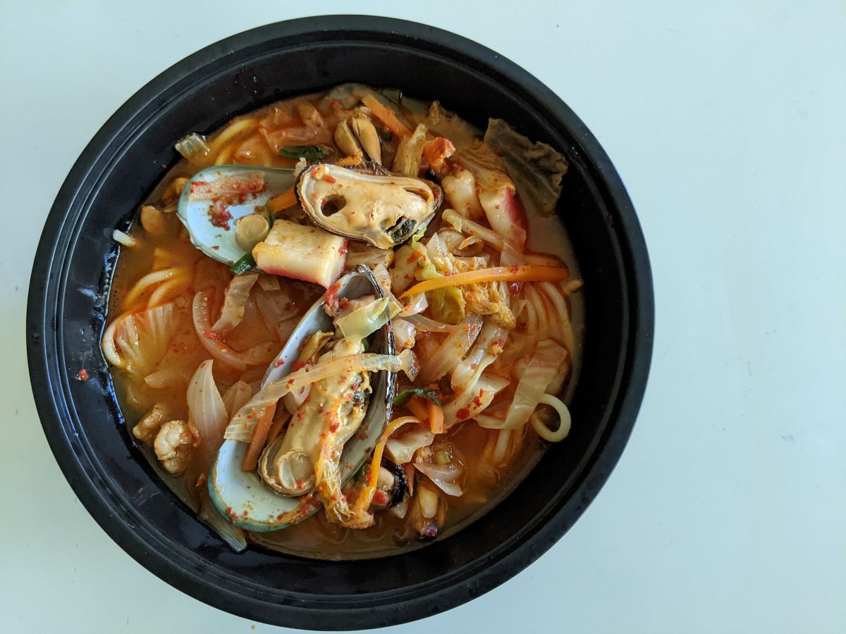 A red seafood noodle soup with large mussel shells in a black plastic bowl. The bowl sits atop a white table. Photo by Tias Paul.