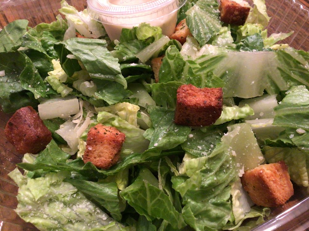 A slightly angled view of the individual Caesar salad shows fresh, chopped romaine and iceberg lettuce, a sprinkle of parmesan cheese, scattered croutons, and a ramekin of creamy Caesar dressing. Photo by Rachael McMillan.
