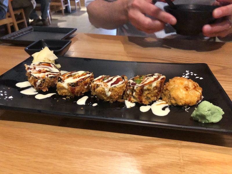 A fried sushi roll from Sakanaya sits on a black rectangular plate with an orange drizzle and an eel sauce drizzle. There is a square soy sauce bowl and a square side plate in the background. Photo by Alyssa Buckley.