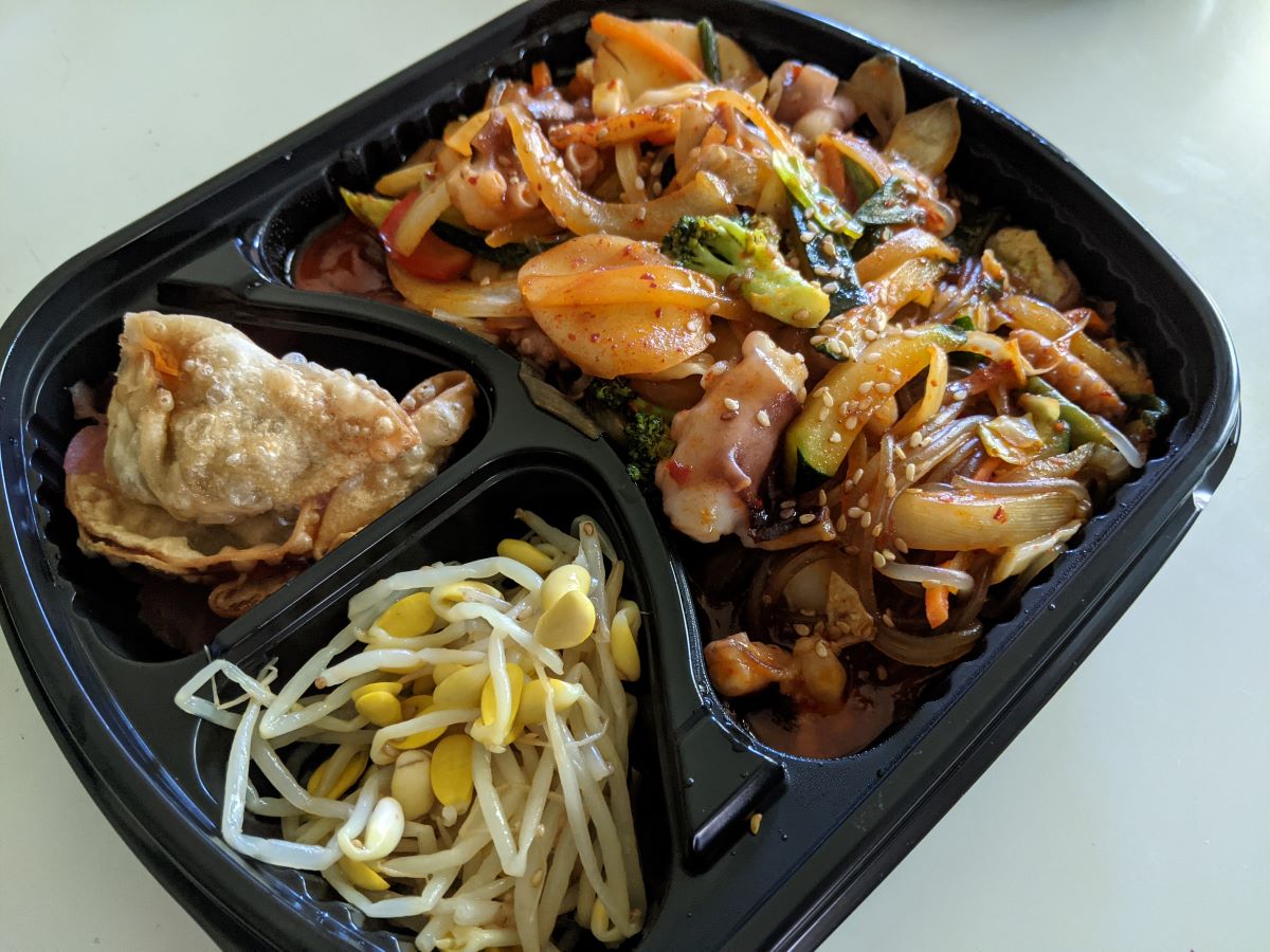 A red-hued mixture of octopus, rice cakes, and vegetables in the large compartment of a black, three-compartment tray. Other compartments feature dumplings and sprouts. The tray is on a white table. Photo by Tias Paul.
