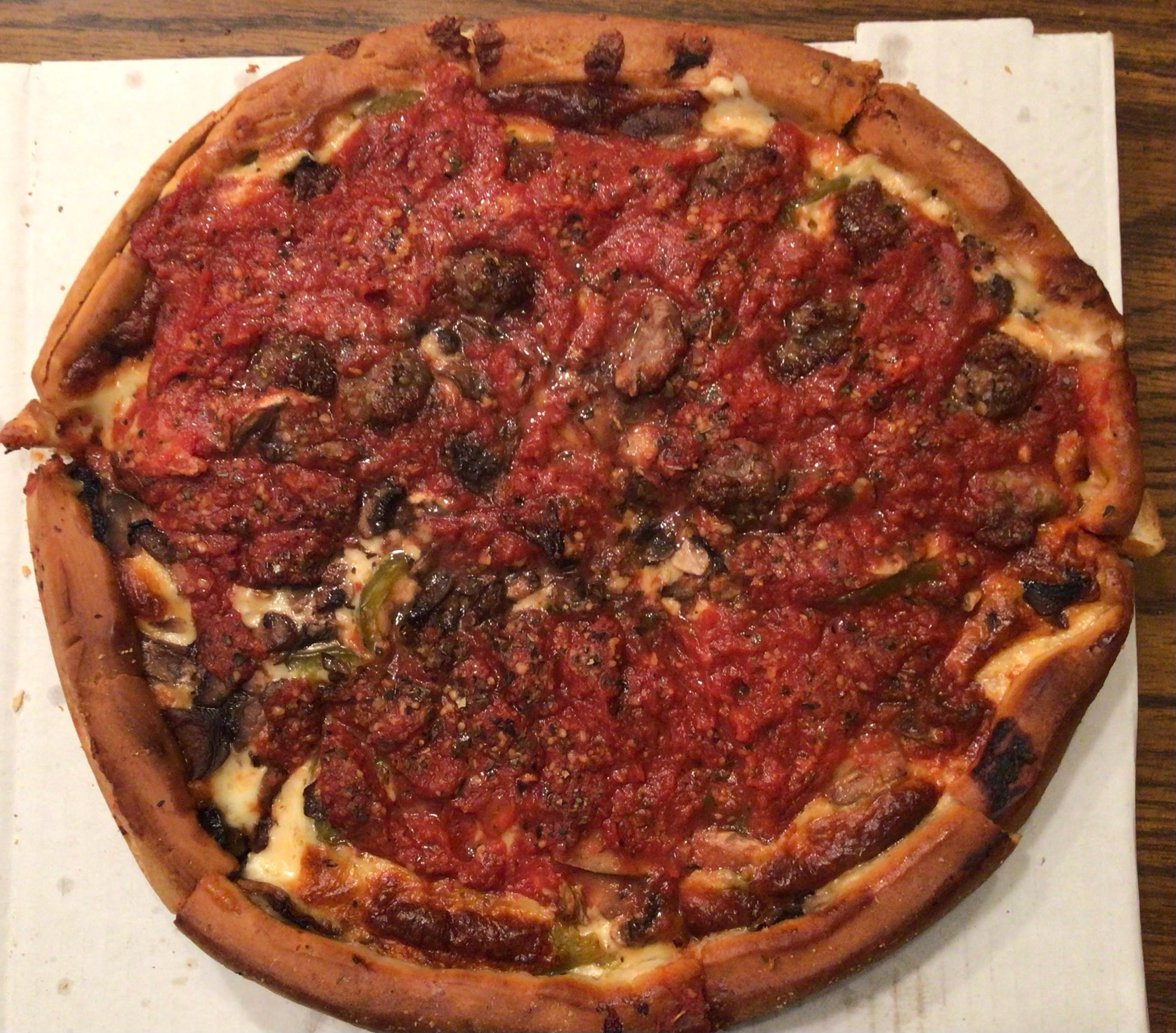 Papa Delâ€™s Sicilian Pan Pizza is shown whole, photographed from above: an approximately two-inch deep crust (darkened slightly at the edges) holds some visible toppings, such as sausage and green pepper. The rich, red pizza sauce covers the top completely. Photo by Rachael McMillan.