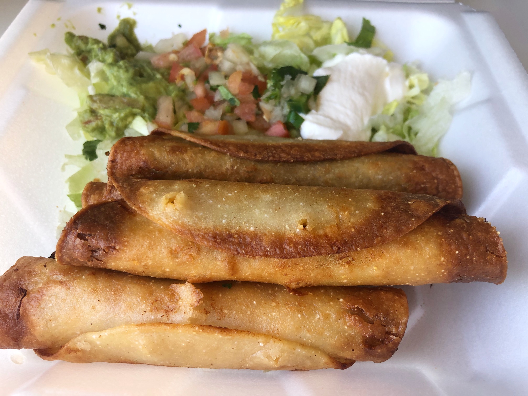 In a styrofoam takeout container, there are three flautas with brown edges from the fryer and in the two dividers behind, there is sour cream, guacamole, pico, and lettuce. Photo by Alyssa Buckley.