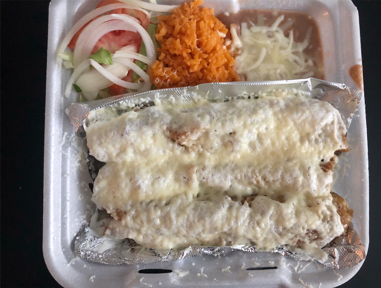 In a styrofoam container, there are three flautas covered in cheese atop tin foil. In the other divider, from the left, there are onion slices, a tomato slice, lettuce, then Mexican rice, and then beans. Photo by Alyssa Buckley.