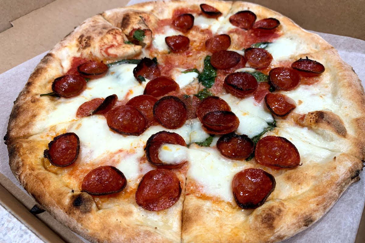 A photo of a small, thin crust pizza on top of parchment paper in a cardboard box. The crust is light brown, the cheese is very white, and the pizza is topped with red pepperoni slices and large pieces of green basil. The pizza is cut into slices. Photo by Megan Friend.