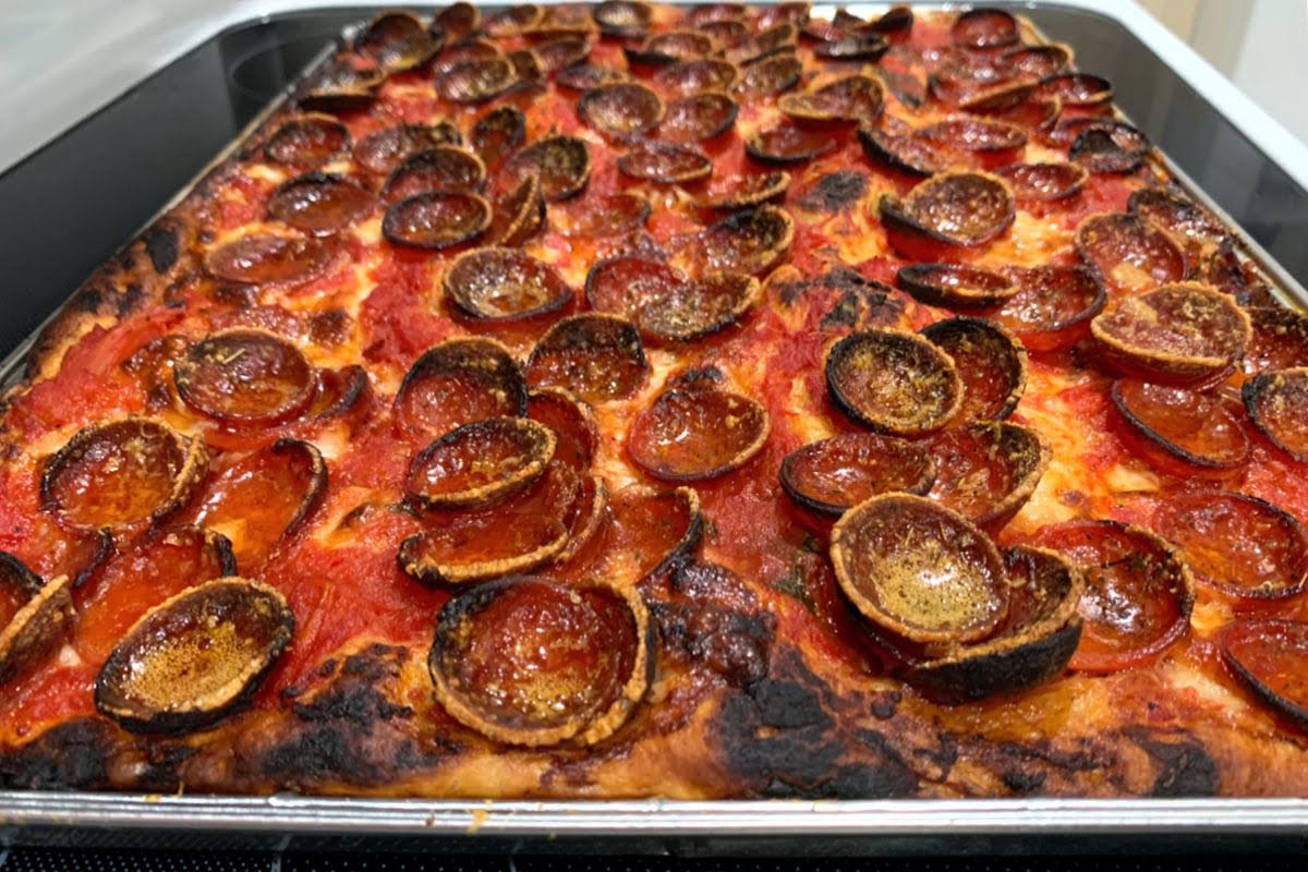 A side-view photo of a large, thick crust Sicilian pizza in a silver baking pan sitting on a stove top. Parts of the crust are charred, and the surface of the pizza is covered in small, cooked pepperoni cups. Some of the pepperonis are also charred. The pizza is uncut. Photo by Megan Friend.