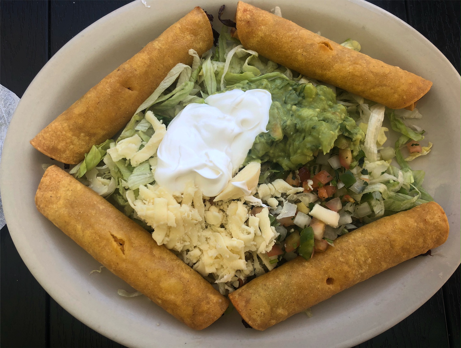 On a white oval plate, there are four flautas in a ring, and inside, there are toppings like sour cream, pico de gallo, grated white cheese, and guacamole. Photo by Alyssa Buckley.